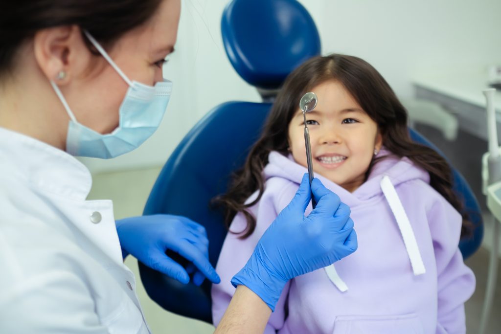 a female child sits in a dentist chair and smiles at the toll the dentist is holding.