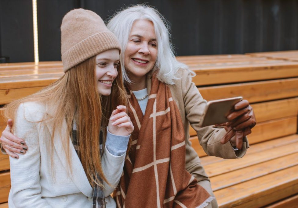 a mature aged woman sites with a young woman as they take a selfie on a phone